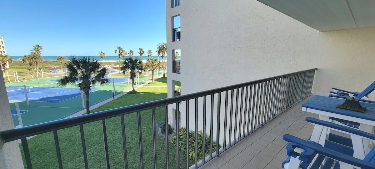 A balcony with a view of a tennis court, palm trees, and a building.