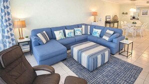 Ein Wohnzimmer mit einem blauen Ecksofa, einem gemusterten Hocker, einem Couchtisch und einem Essbereich mit weißen Stühlen und einem Tisch.