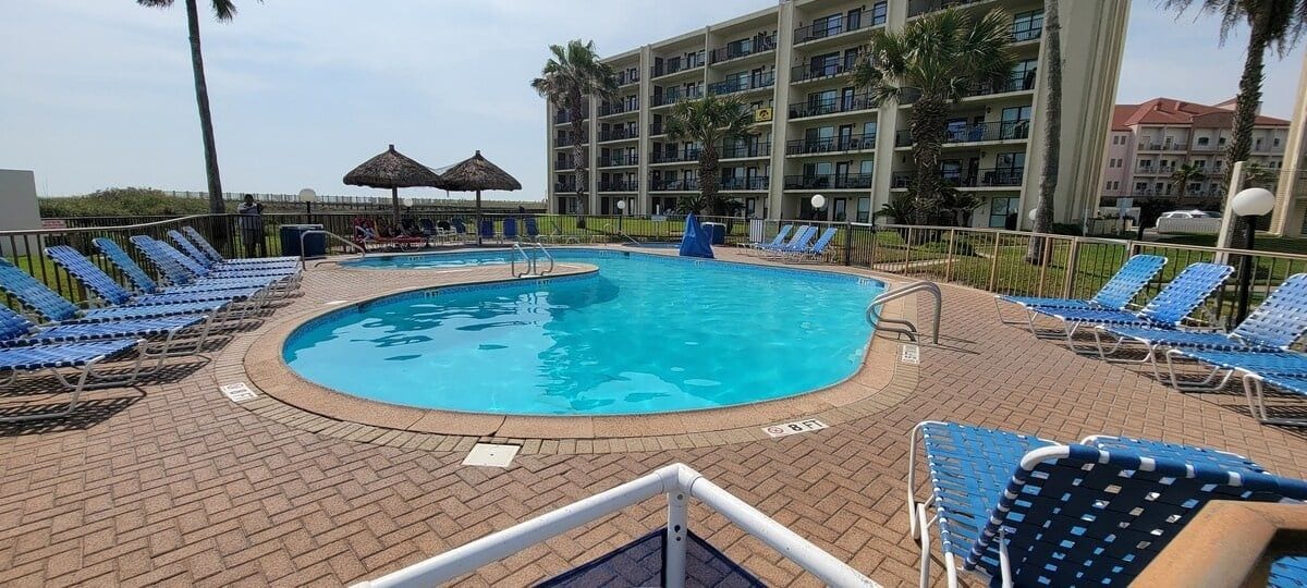 A pool area with lounge chairs, a pool, and a multi-story building in the background.