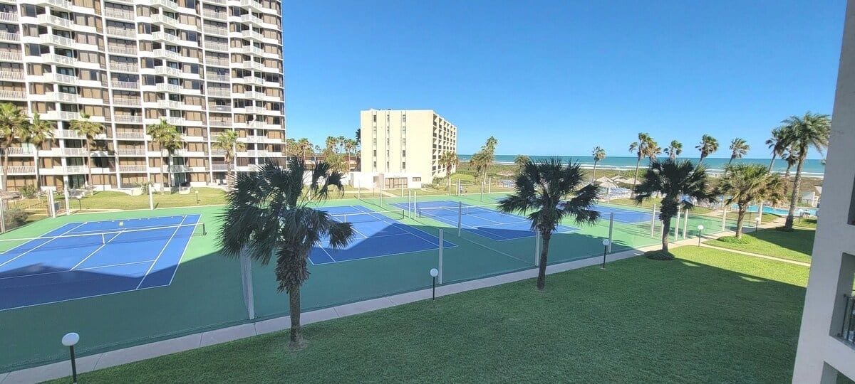 A tennis court area with a view of a tall building and the ocean.