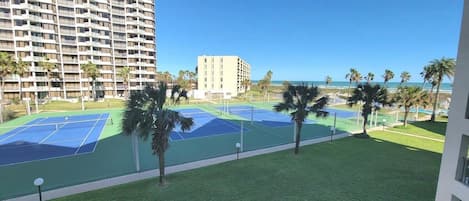 A tennis court area with a view of a tall building and the ocean.