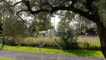 A hammock swing under a tree in a grassy area with a road and buildings in the background.