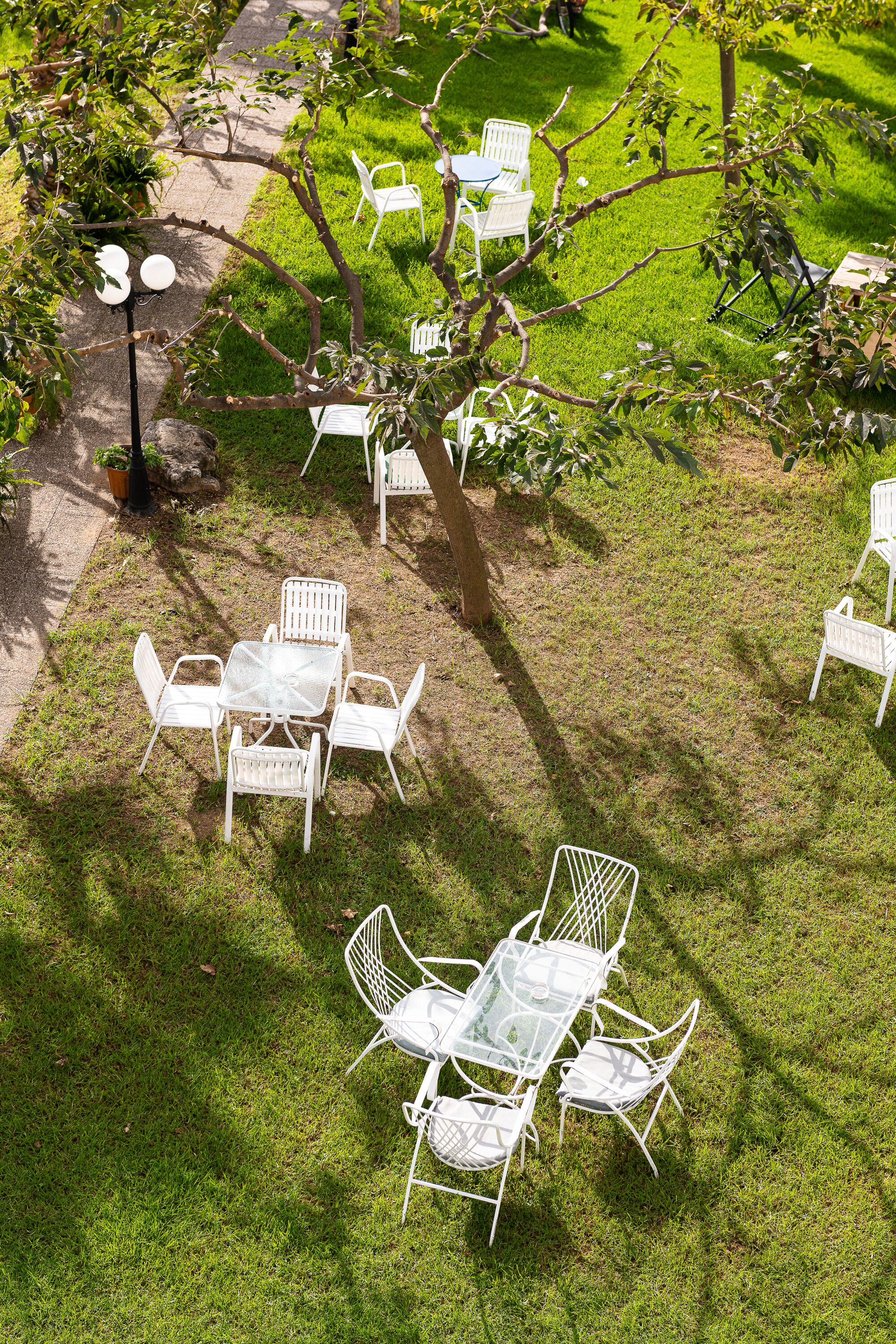 A patio with white metal chairs and tables, surrounded by green grass and trees.