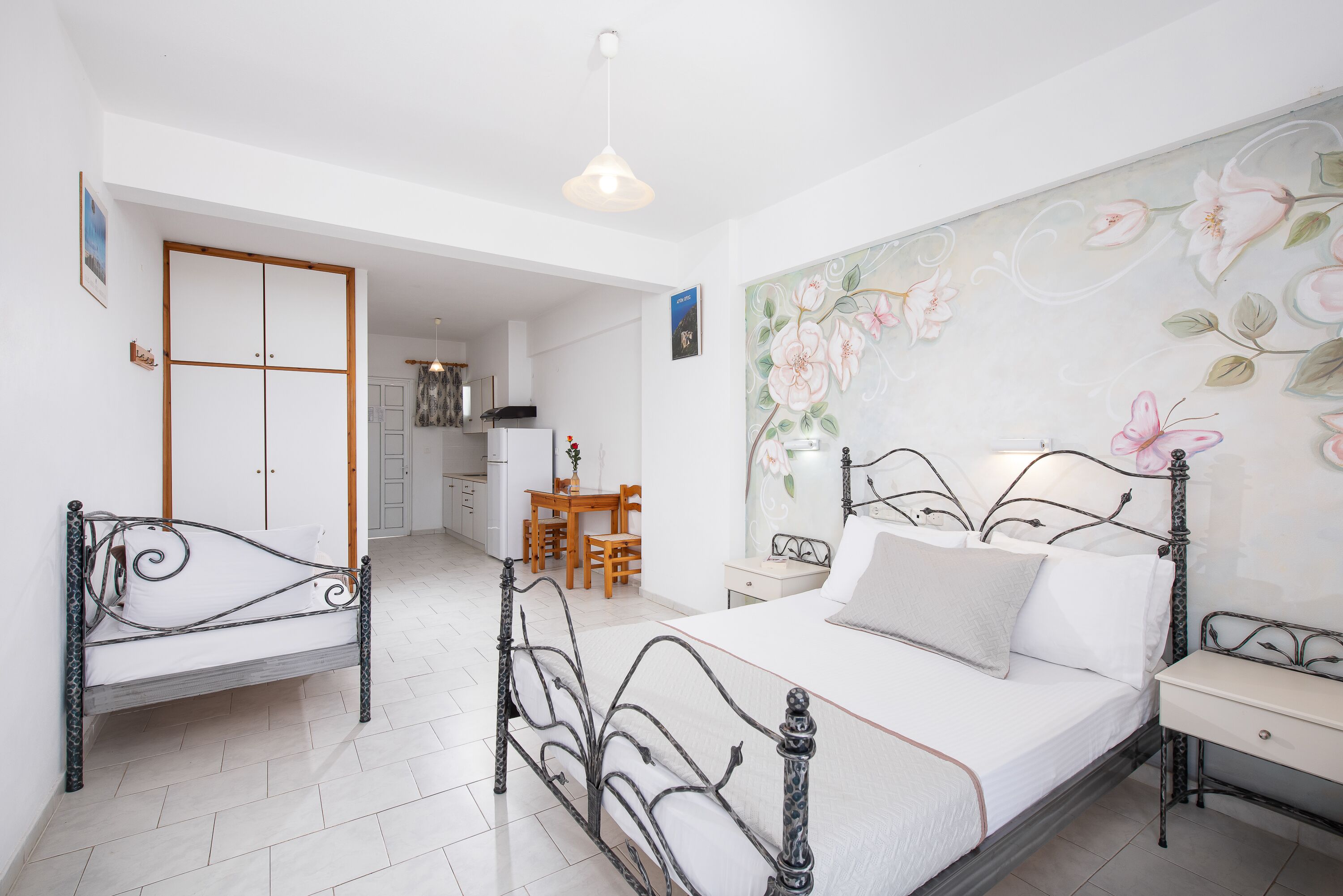 A bedroom with a bed, bedside tables, a desk, and a chair. There is a door leading to a kitchen and a dining area.