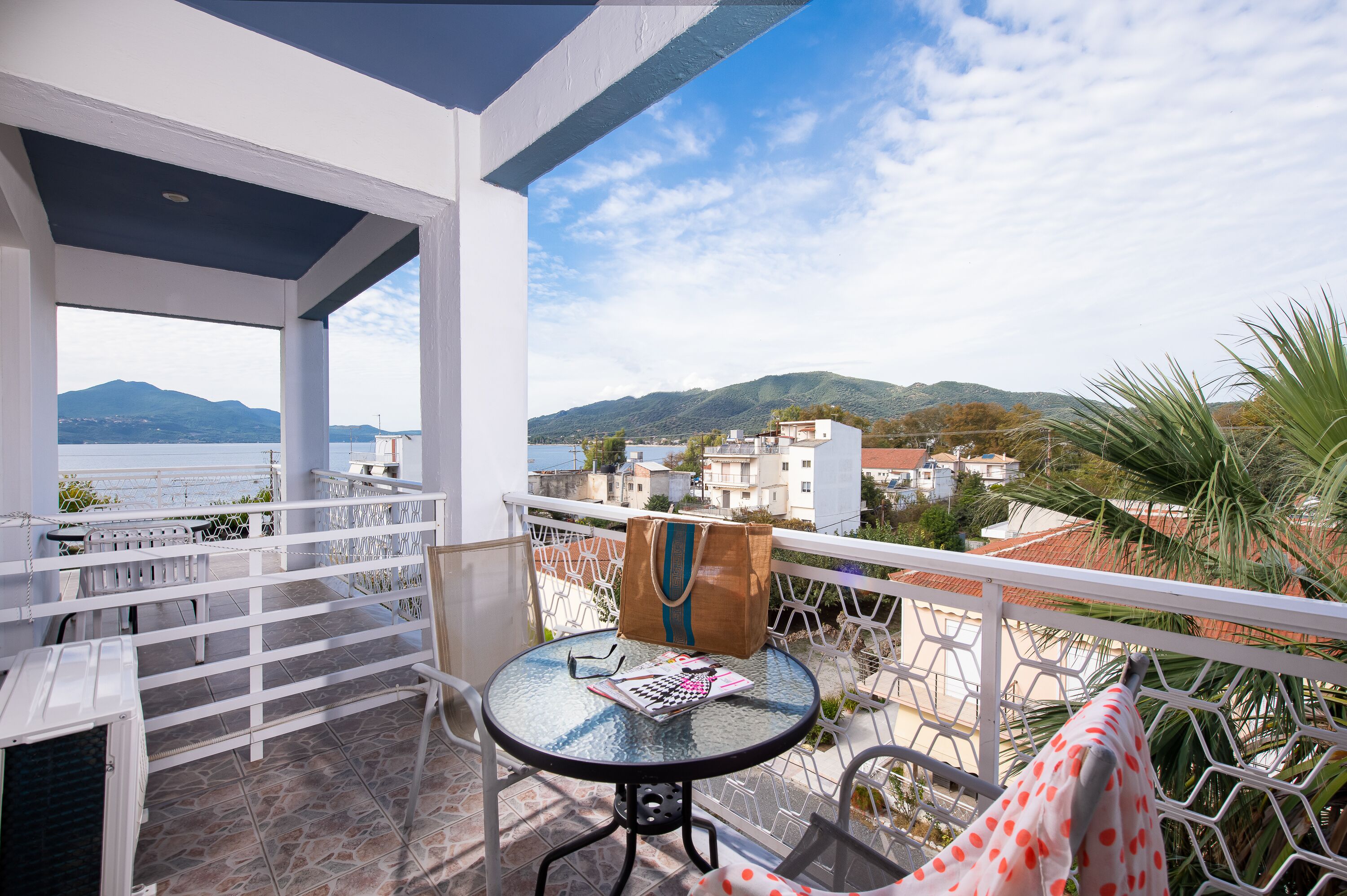 Un balcon avec une table, des chaises, un sac et une vue sur la mer et les montagnes.