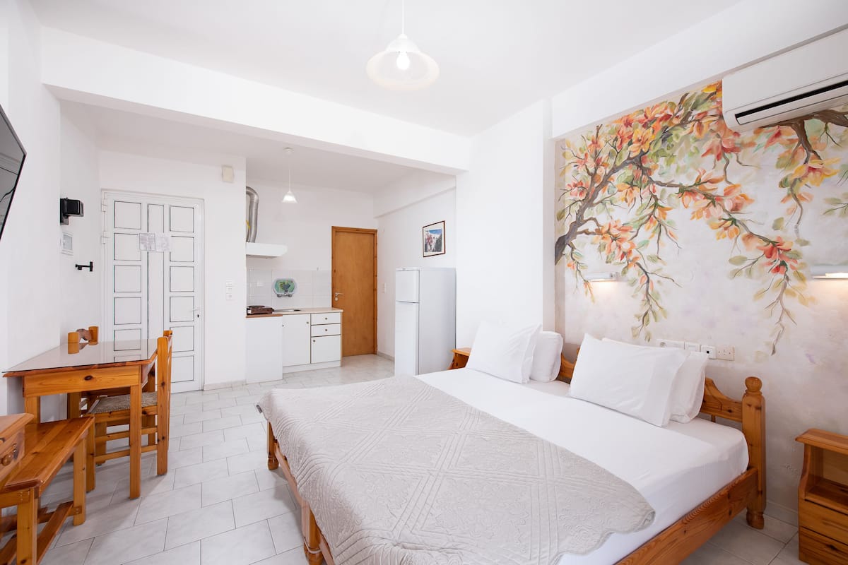A bedroom with a large bed, a wooden nightstand, a desk with a chair, and a kitchenette in the background.