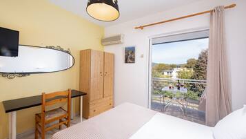 Un dormitorio con cama, escritorio, silla, armario, balcón con mesa y sillas, y vistas al exterior.