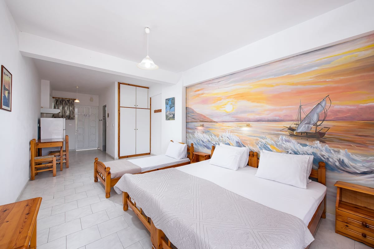 A room with two beds, a desk, and a chair. There is a painting of a sunset on the wall.