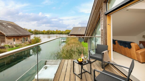 Ein moderner Balkon mit Glasgeländer und Blick auf einen Fluss, mit Holzdeck und einer Sitzecke mit Sofa und Sesseln.