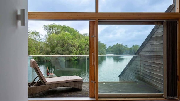 A balcony with a view of a lake and forest, a lounge chair, and a glass door.
