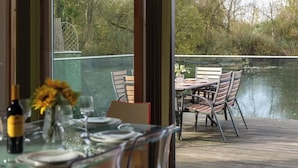 A dining table set with plates, glasses, and a bottle, overlooking a calm body of water and a wooden deck.