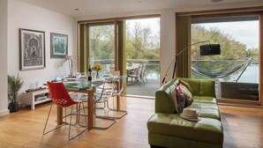 A modern living room with a green sofa, a dining area with clear chairs, and large windows overlooking a body of water.