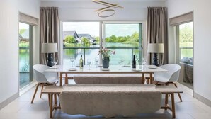 A dining area with a table set for four, large windows with curtains, and a view of a lake and house.