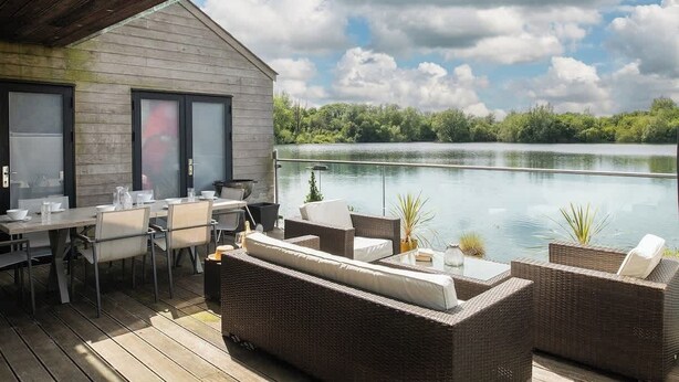 An outdoor deck with a dining area, wicker furniture, and a view of a lake. - Egret Lodge (South Cerney)