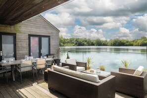 An outdoor deck with a dining area, wicker furniture, and a view of a lake. - Egret Lodge (South Cerney)