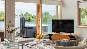 A modern living room with a large TV, a sofa, a coffee table, and a view of a boat through large windows. - Egret Lodge (South Cerney)