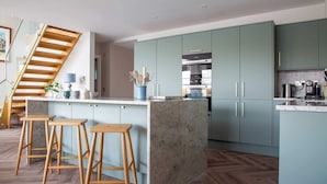 A modern kitchen with a central island, wooden stools, and a staircase. - Egret Lodge (South Cerney)