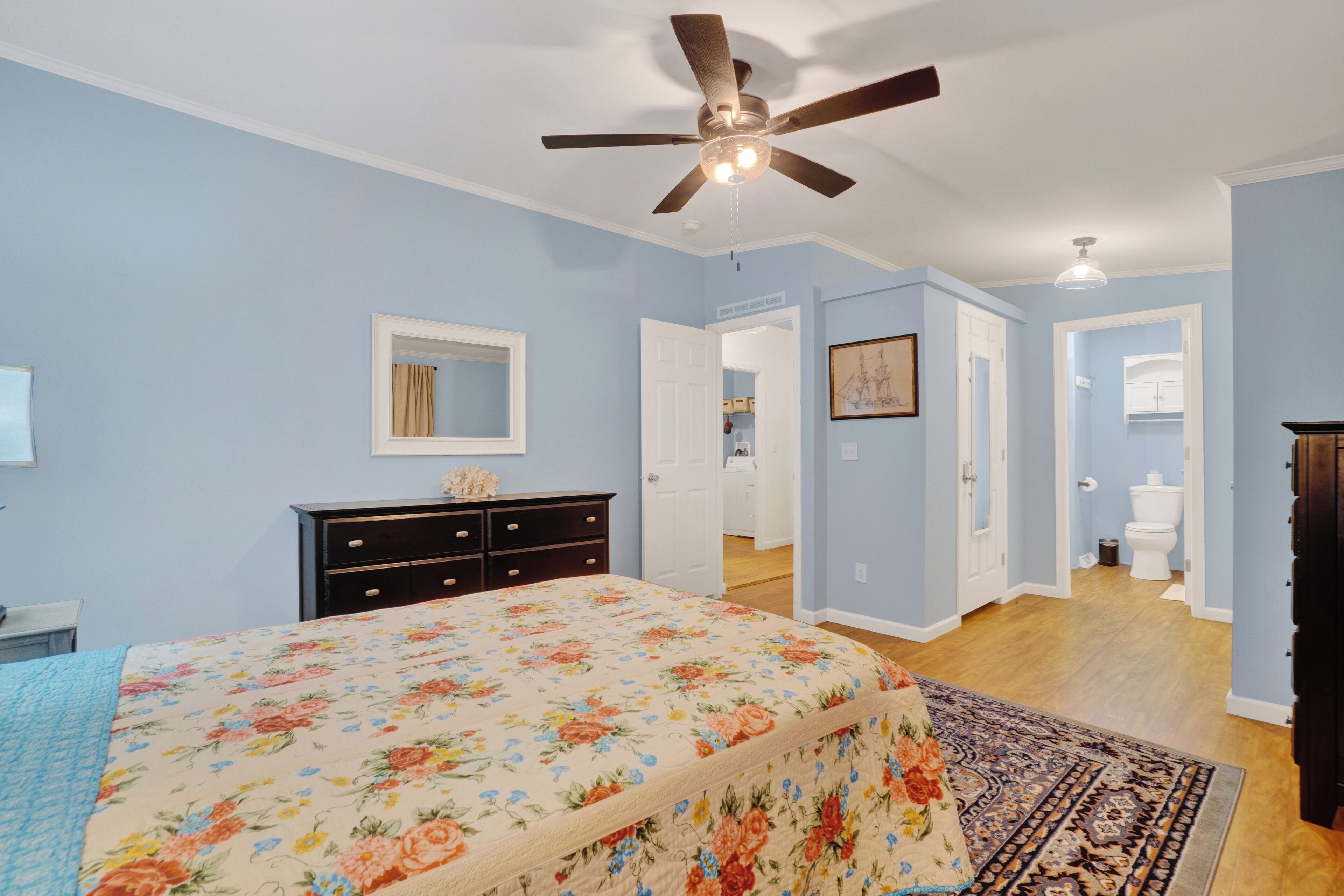 A bedroom with a bed, dresser, ceiling fan, and a hallway leading to a bathroom.