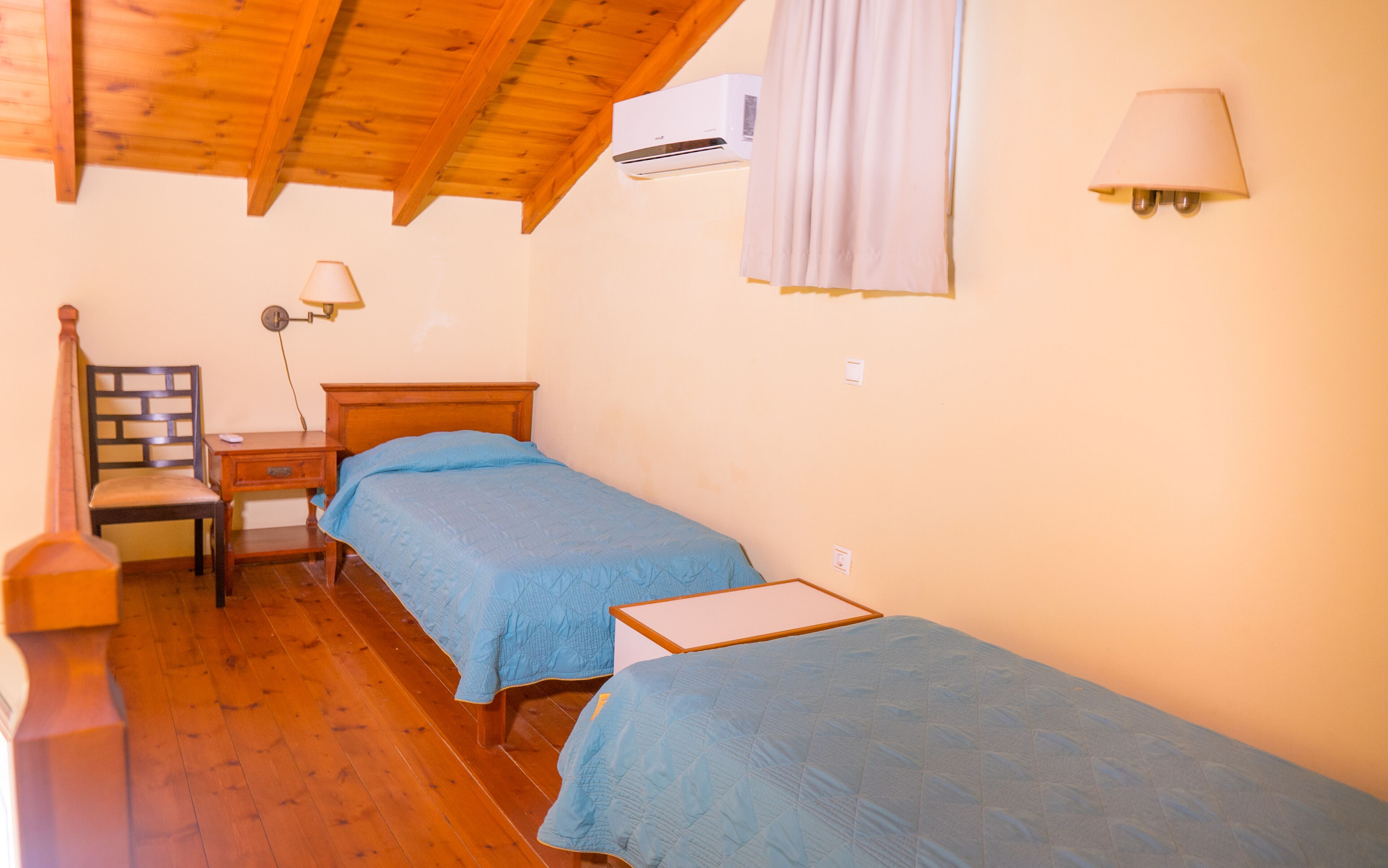 A room with two beds, a wooden ceiling, an air conditioning unit, a lamp, and a chair.