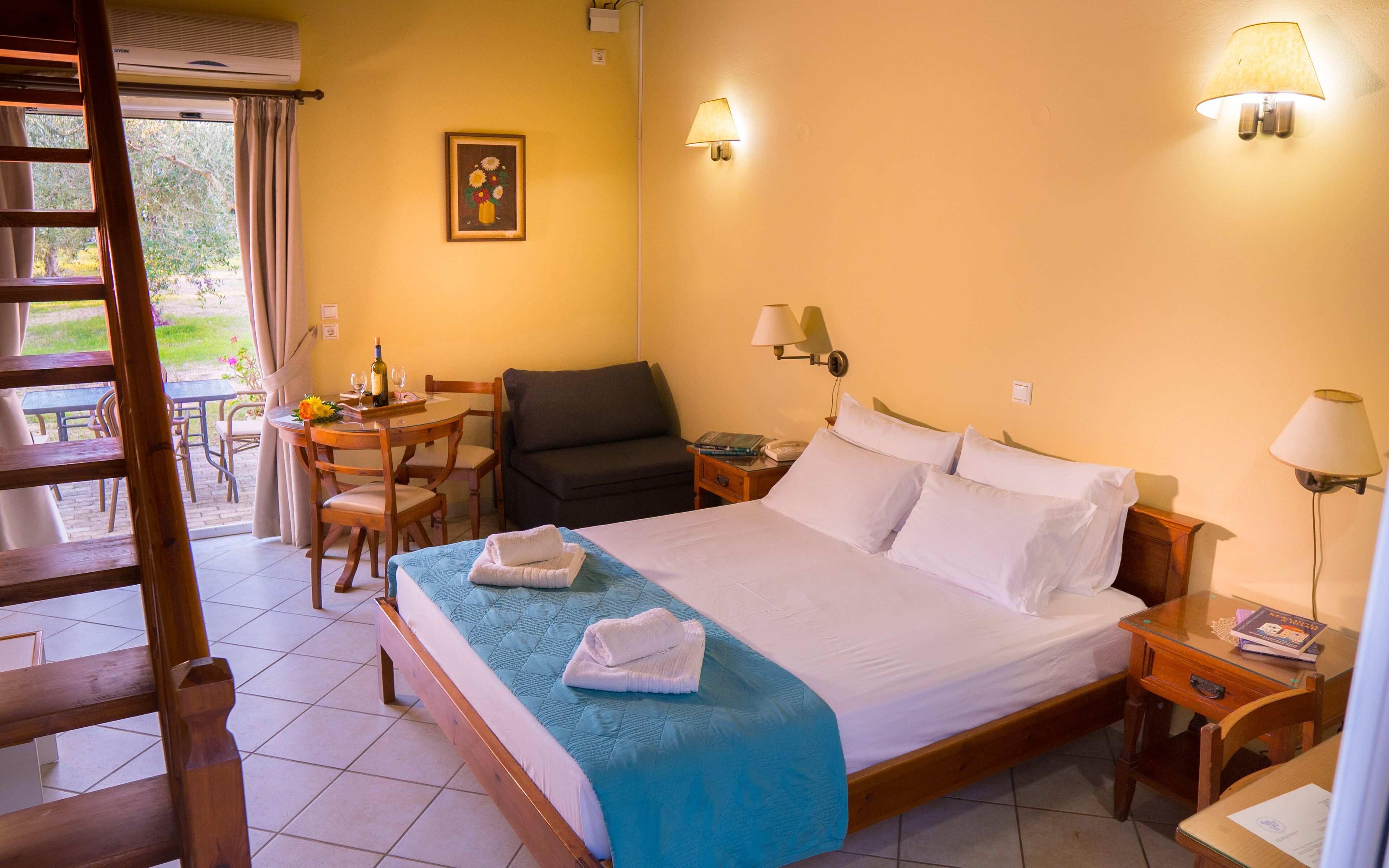 A hotel room with a bed, bedside tables, a desk with chairs, a sofa, and a window with curtains.