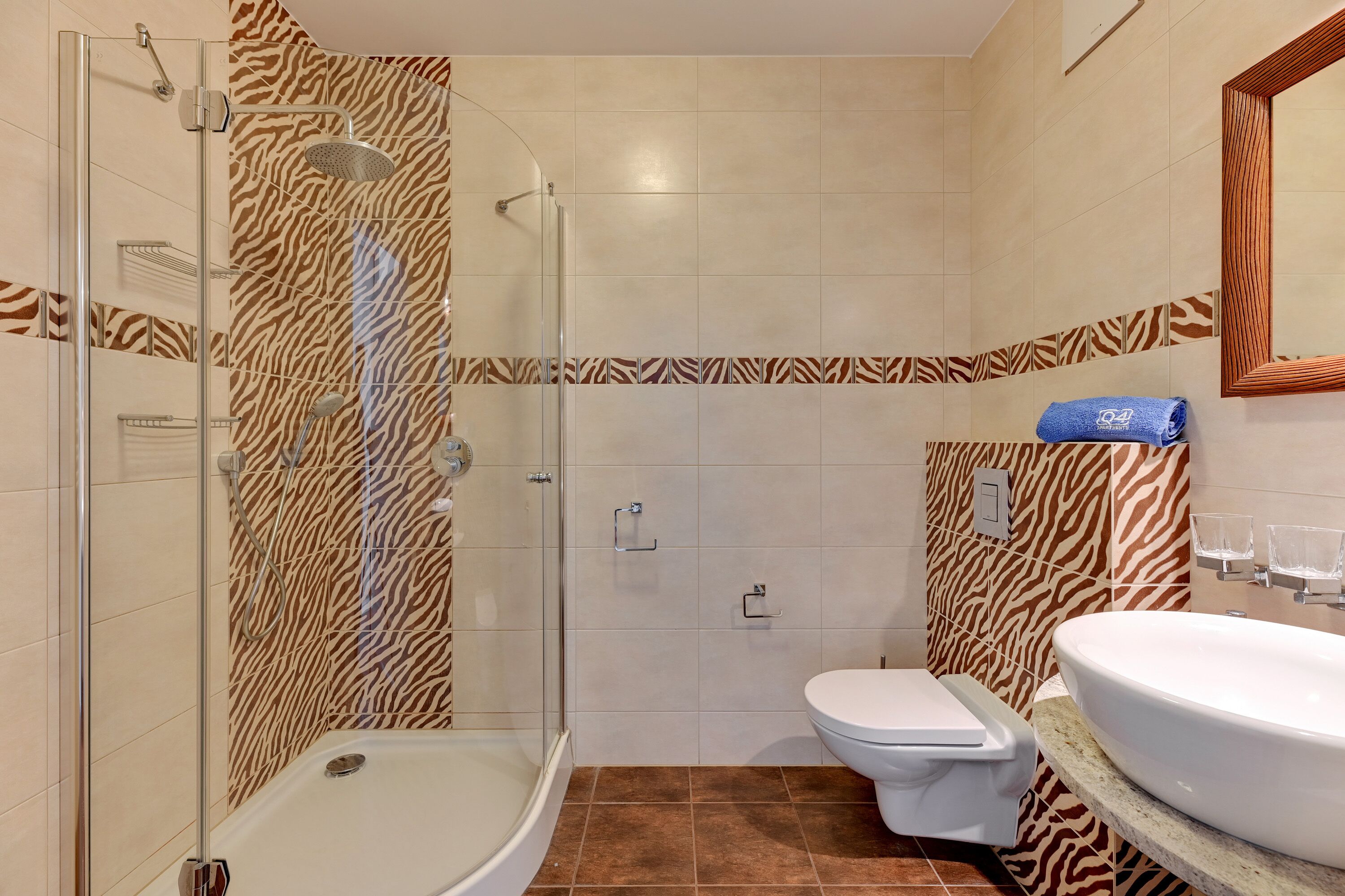 Ein Badezimmer mit einer gläsernen Duschkabine, einer weißen Toilette und einem Waschbecken mit einer waschbärenmusterigen Fliesenbordüre.