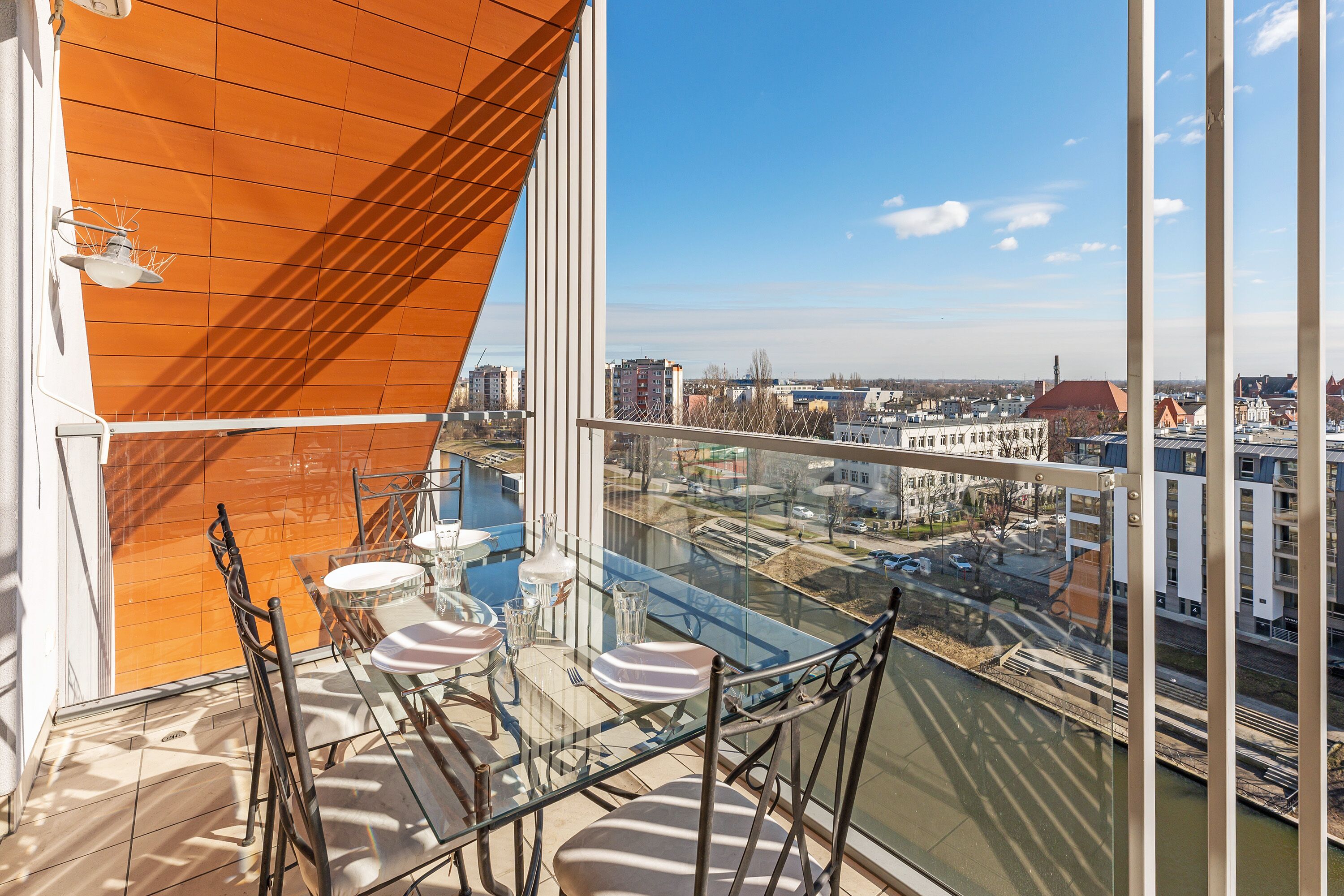 A high-rise balcony with a glass table and chairs, offering a view of a cityscape and a river.
