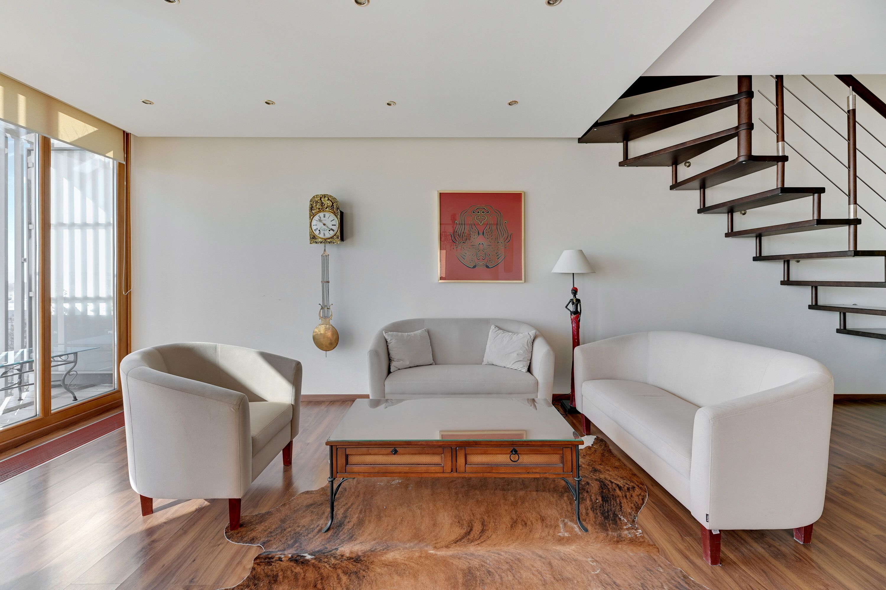 Een moderne woonkamer met een bank, fauteuil en salontafel, een trap en een groot raam.