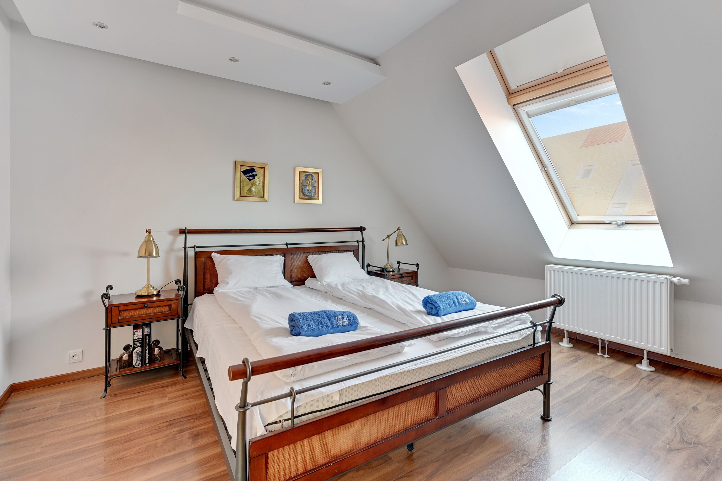 Una camera da letto con soffitto spiovente, un lucernario, un letto con lenzuola bianche, due asciugamani blu, un comodino con una lampada e un termosifone.
