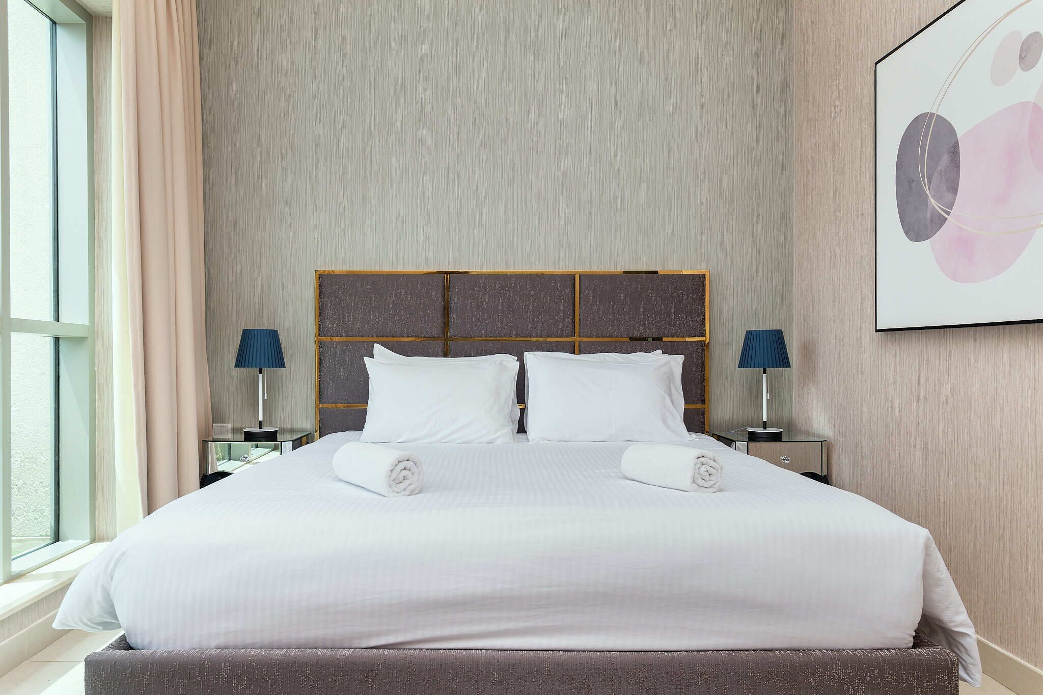 A neatly made bed with white linens and two pillows, flanked by two bedside tables with lamps, in a room with a large window and a framed abstract artwork on the wall.