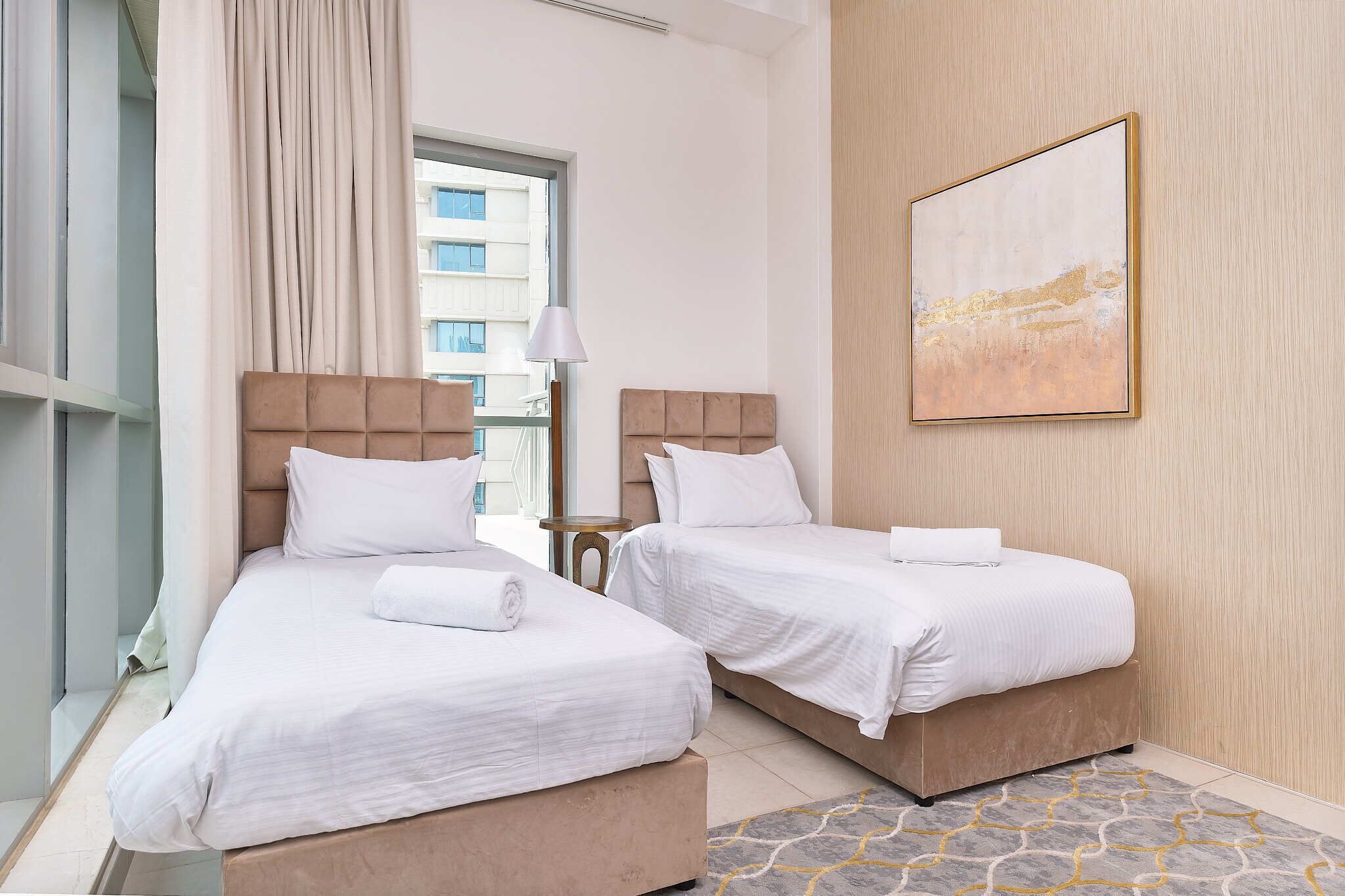 A hotel room with two beds, a large window, a painting on the wall, and a patterned rug.