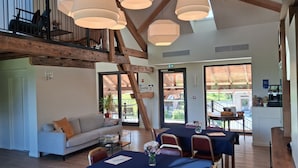 Una habitación amplia con techo alto, vigas de madera, un comedor con mesas y sillas, un sofá y una escalera que sube a un nivel superior.
