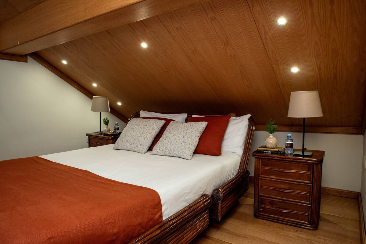 A bedroom with a sloped ceiling, a bed with white linens, two nightstands with lamps, and a small plant on one nightstand.