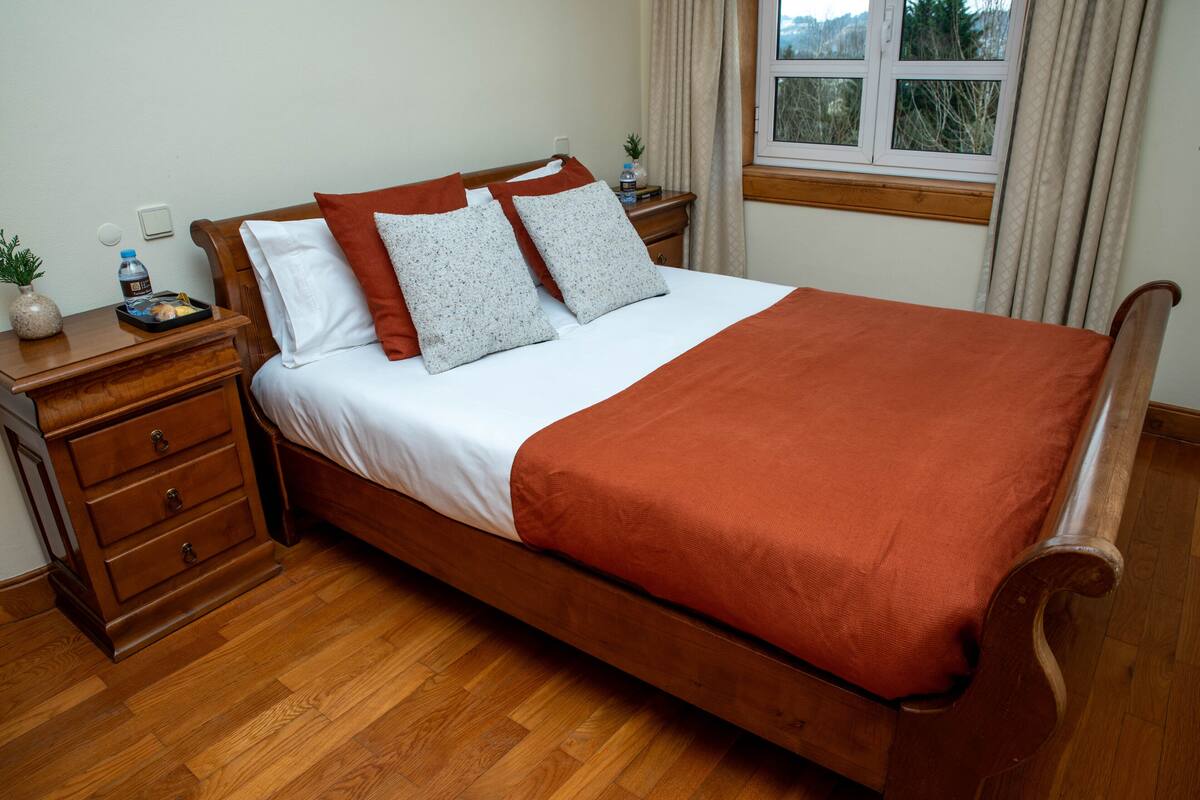 A neatly made bed with a wooden frame, white bedding, and a red blanket. There is a wooden nightstand with a lamp and a small plant beside the bed. A window with curtains is visible in the background.
