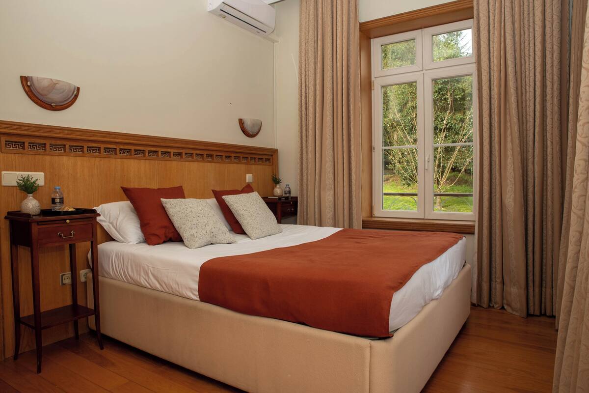 A neatly made bed with a brown blanket, a bedside table with a lamp, and a window with curtains.