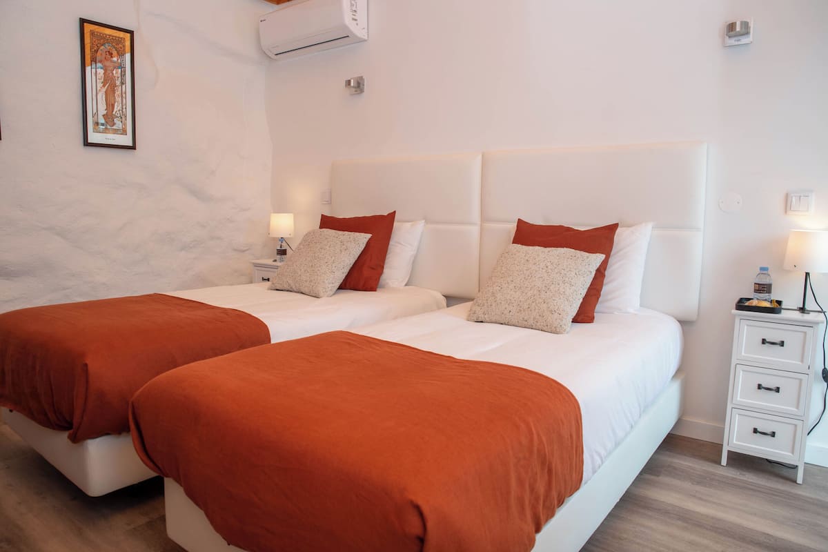 A hotel room with two beds, each with white bedding and orange throw pillows. There is a white nightstand with a lamp and a small table with a bottle.