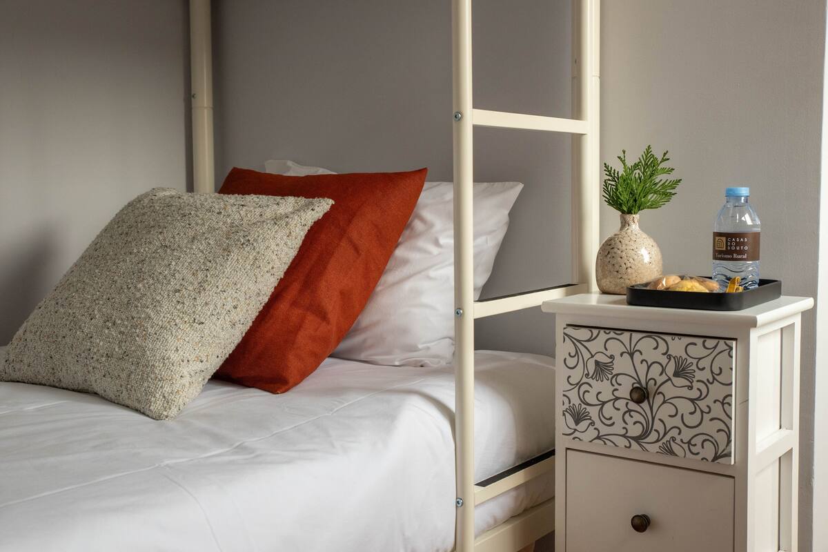 A bunk bed with white bedding and a red pillow, a small table with a vase and a water bottle, and a decorative metal drawer.