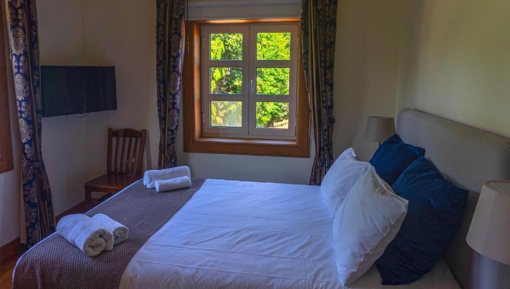 Una cama bien hecha con sábanas blancas, una mesita de noche de madera, una silla y una ventana con vistas a la vegetación.
