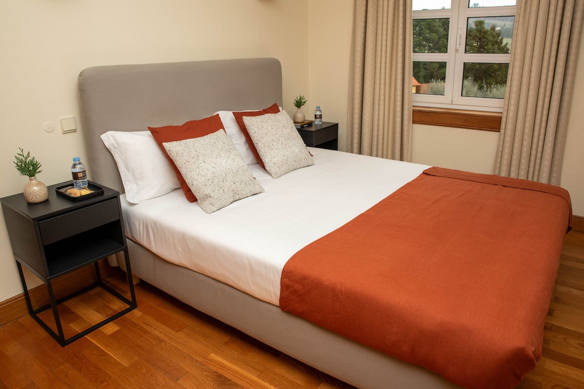 A neatly made bed with a grey headboard, two pillows, and a brown throw blanket. There is a small table with a water bottle and a plant beside the bed.