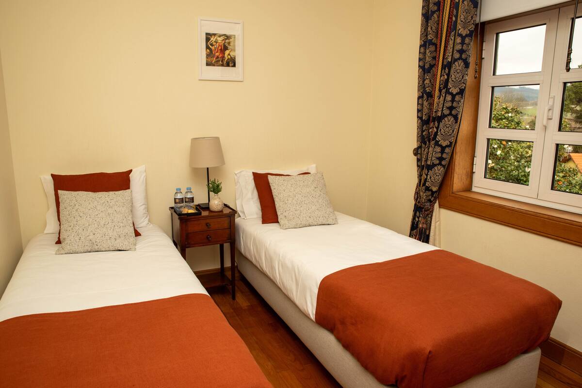 A hotel room with two beds, a nightstand with a lamp, a framed picture on the wall, and a window with a view of greenery.