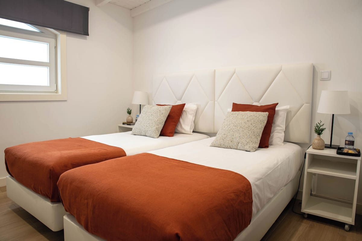 A hotel room with two beds, each with a white headboard and brown bedding. There are two bedside tables with lamps and a small plant on one.