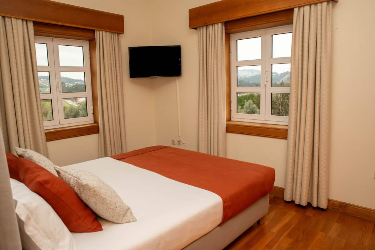 A hotel room with a bed, two windows with curtains, a flat-screen TV mounted on the wall, and a view of greenery outside.