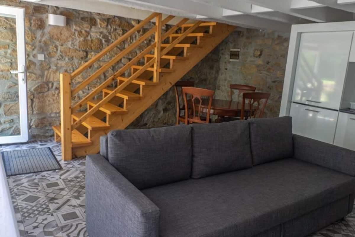 A modern living room with a grey sofa, a wooden staircase, and a kitchen area.