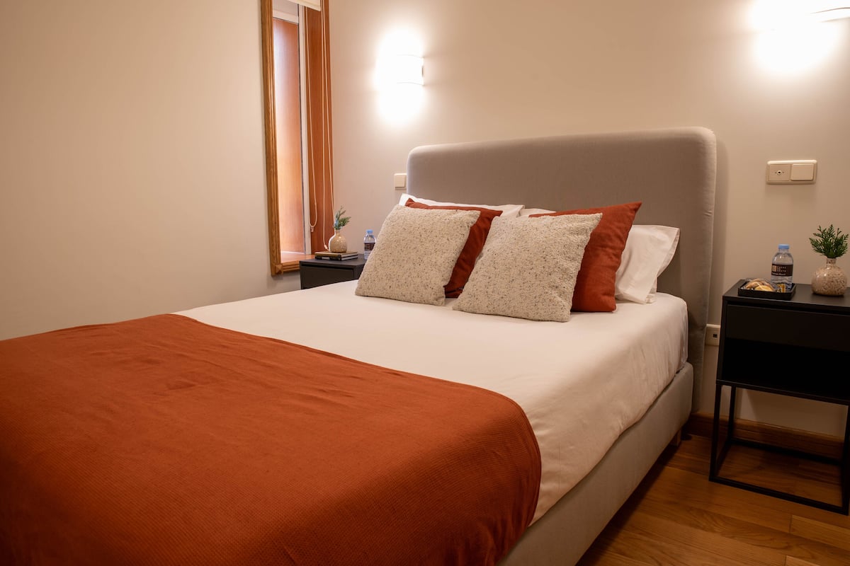 A neatly made bed with a white and orange bedspread, a grey headboard, and two bedside tables with lamps and a water bottle.