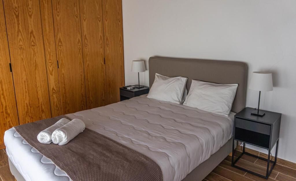 Una cama bien tendida con una manta marrón y dos almohadas, flanqueada por dos mesitas de noche con lámparas, en una habitación con paneles de madera en una pared.