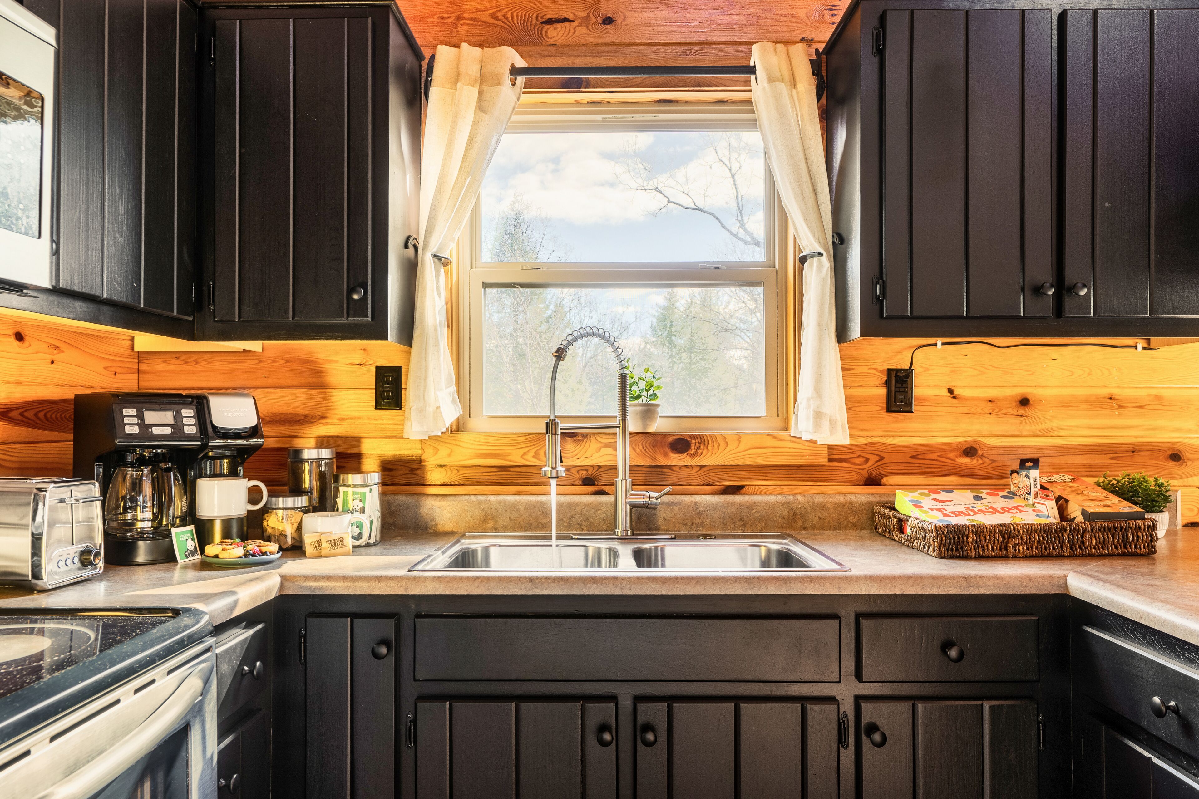 Una cocina con armarios de madera oscura, un lavamanos de acero inoxidable y una ventana con cortinas.
