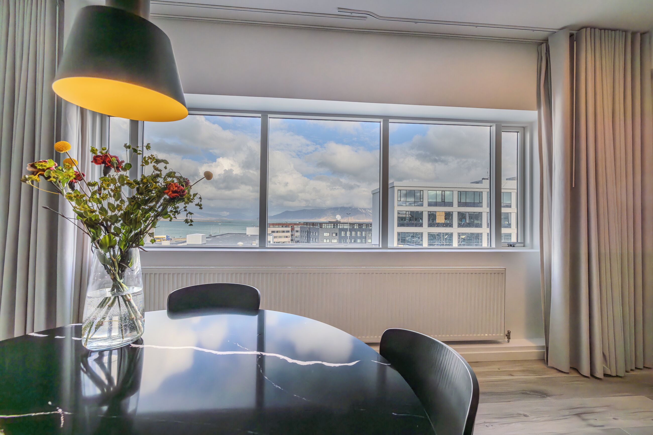 A modern dining area with a round table, chairs, a vase with flowers, and a large window offering a view of buildings and mountains.