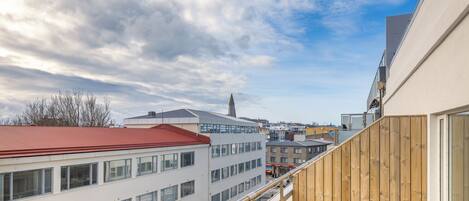 A modern building with a red roof, a wooden balcony railing, and a view of other buildings under a partly cloudy sky.