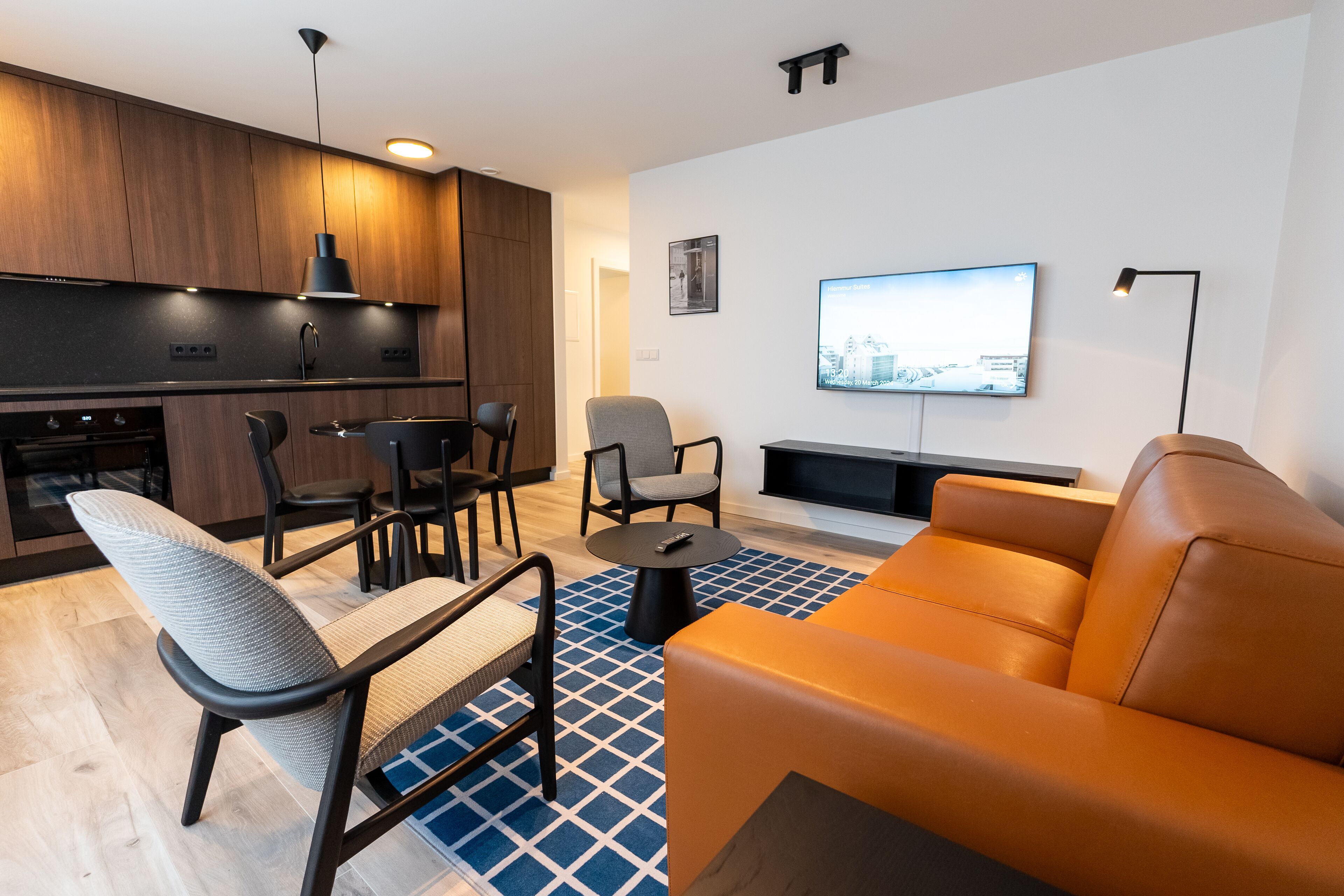 A modern living room with a sofa, chairs, a TV, and a kitchen area with wooden cabinets and a dining table.