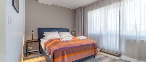 Habitación de hotel moderna con una cama grande, una mesita de noche, una lámpara y vistas al exterior a través de cortinas translúcidas.
