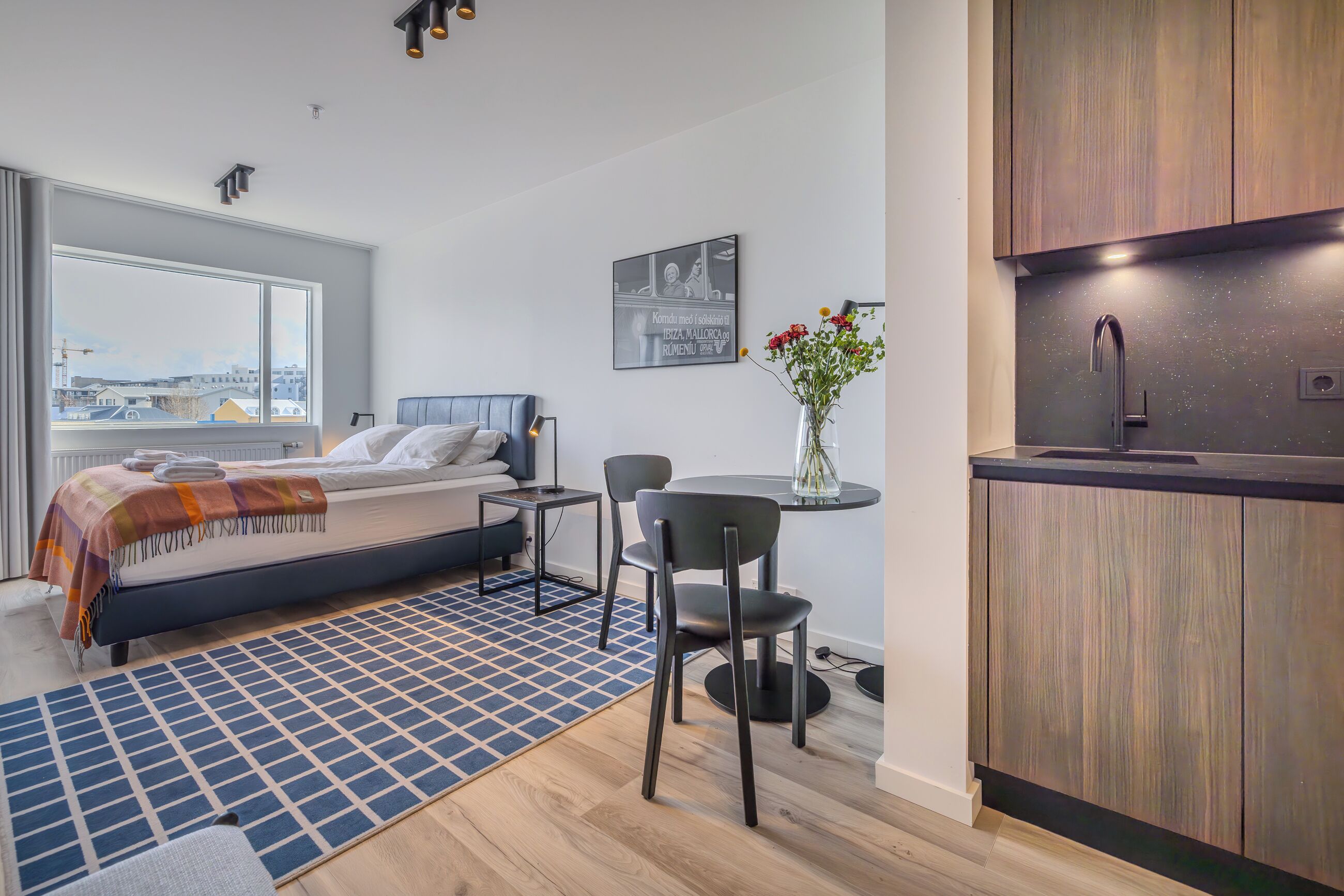 A modern bedroom with a large window, a bed, a bedside table, and a small dining area with a table and chairs.
